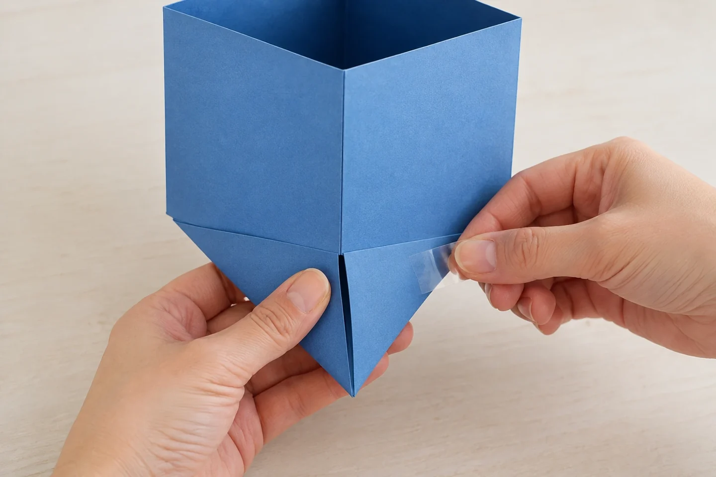 Hands closing the four triangular lower flaps of a square cardstock tube with short exterior tape joins; open top makes the construction visible